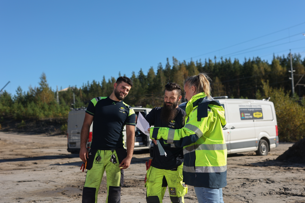1 kvinna och 2 män i varselkläder i bakgrunden syns två arbetsfordon 