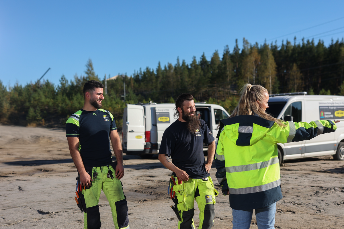 Tre personer i varselkläder med arbetsfordon i bakgrunden 