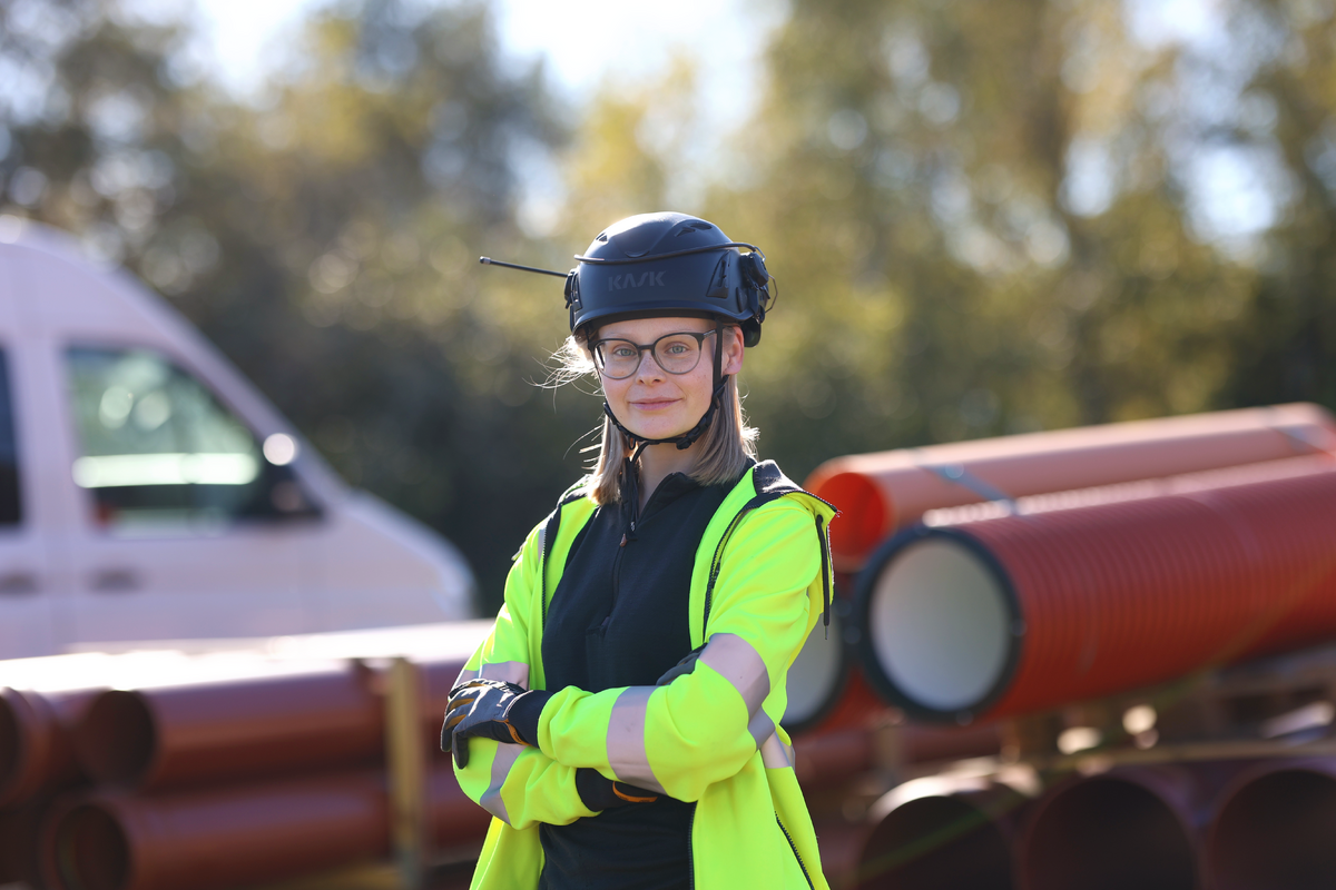 Kvinna i varselkläder och bygghjälm. I bakgrunden syns vattenrör 