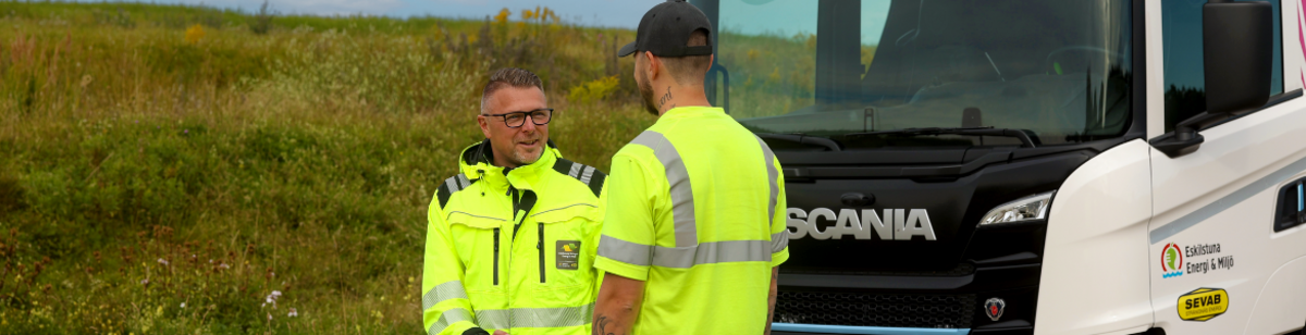 Två män med varselkläder och en lastbil i bakgrunden 