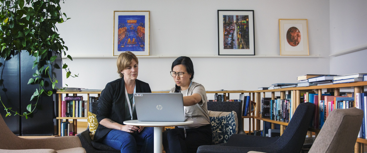 Två personer sitter vid ett bord och tittar på en laptop skärm. I Bakgrunden finns låga bokhyllor och en grön växt. På väggen sitter tre tavlor. 