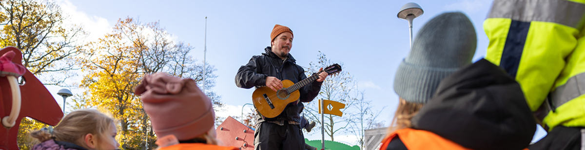 Förskolepersonal som spelar gitarr och sjunger med barnen utomhus.