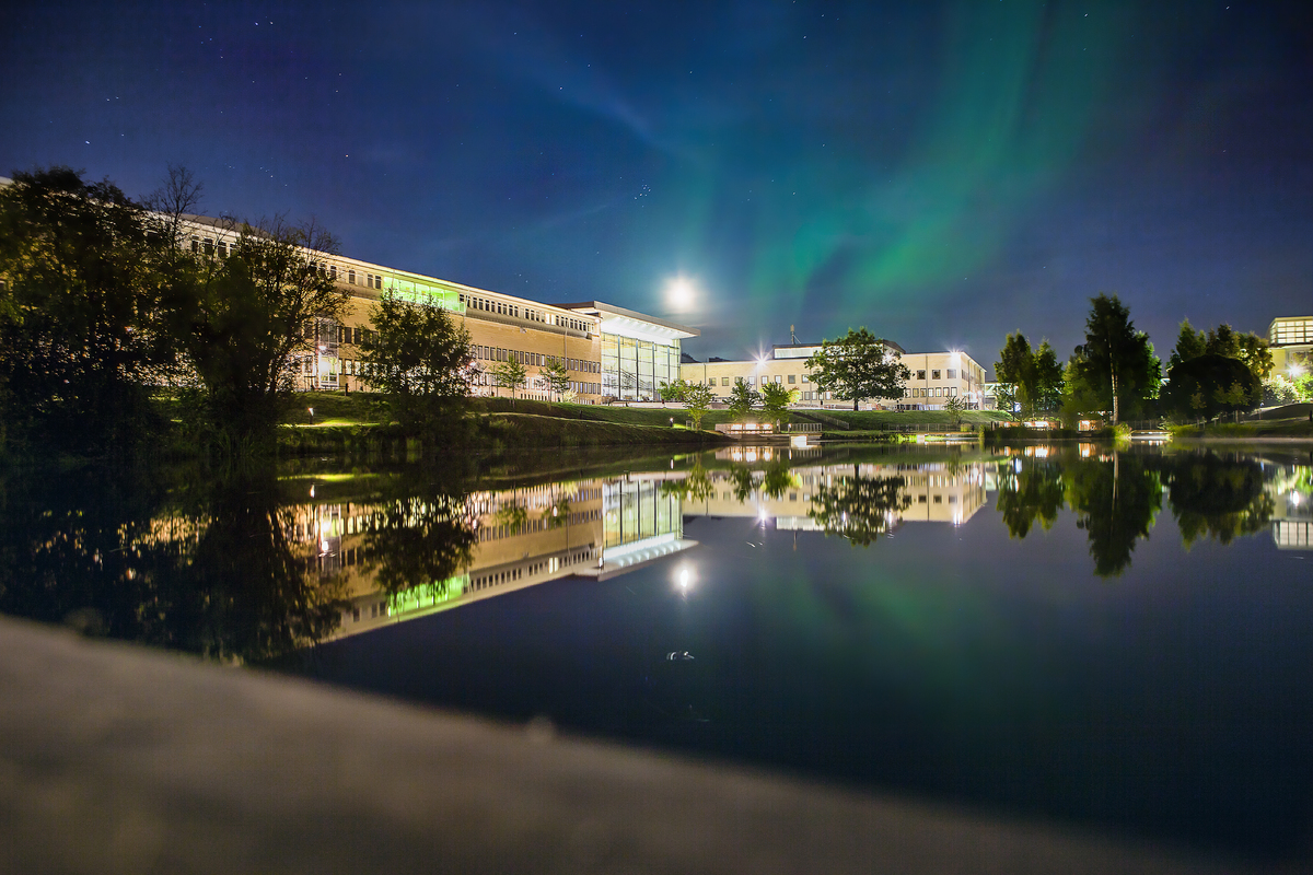Bilden visar ett norrsken över dammen på campus. Lamporna från byggnaderna avspeglar sig vackert i dammen.