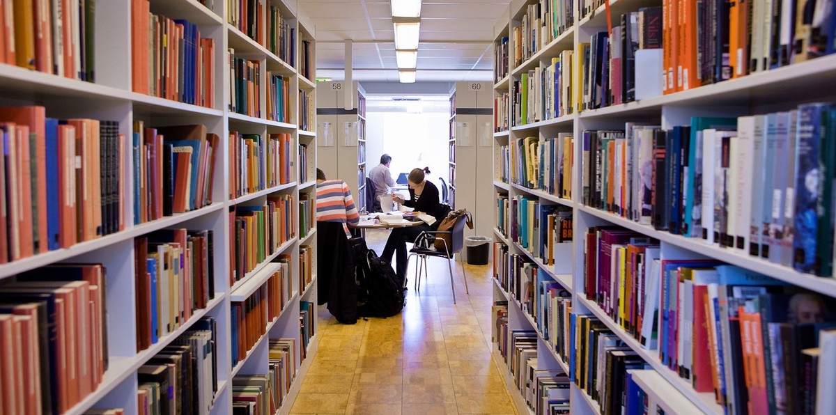 Bilden visar ett bibliotek med flera bokhyllor med böcker i. I mitten av bilden sitter människor runt ett bord och läser.