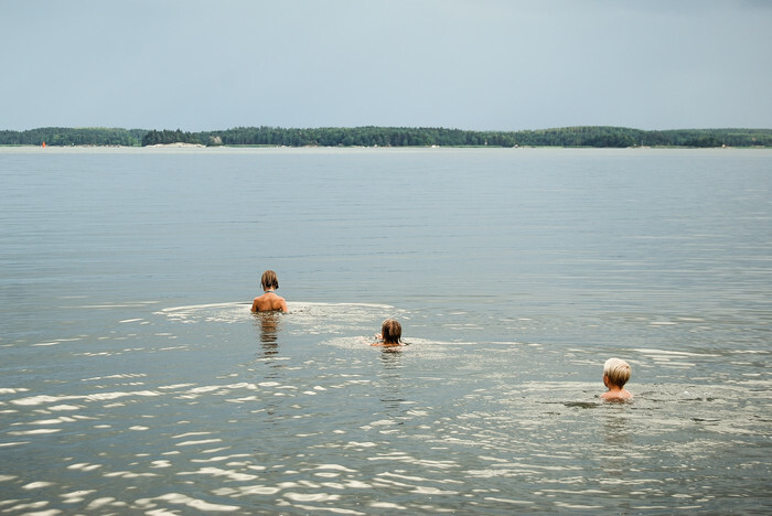 Tre badande i sjö en varm sommardag. Solglitter speglas i vattnet. 