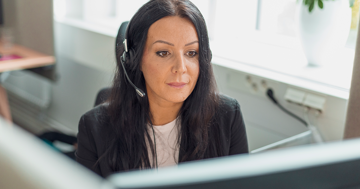 Kvinna sitter framför dator med headset och tittar in i skärmen.