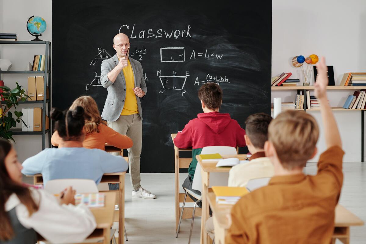 En bild på elever och en lärare i ett klassrum. Läraren har ställt en fråga och en elev räcker upp handen. 