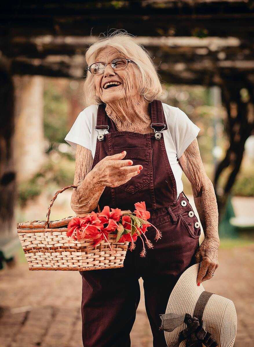 En leende äldre kvinna som står med en korg med nyplockade blommor. 