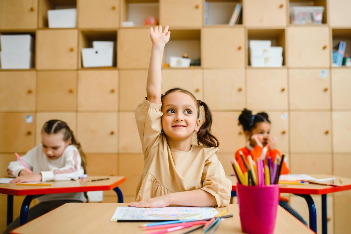 Ett klassrum där vi ser tre flickor, en flicka räcker upp handen och ser glad ut. 