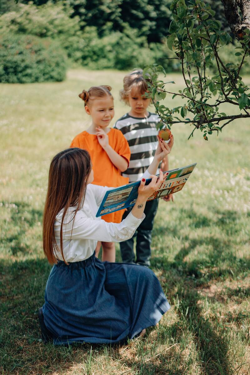 En bild på en kvinna och två elever som är ute i naturen och tittar tillsammans i en bok 