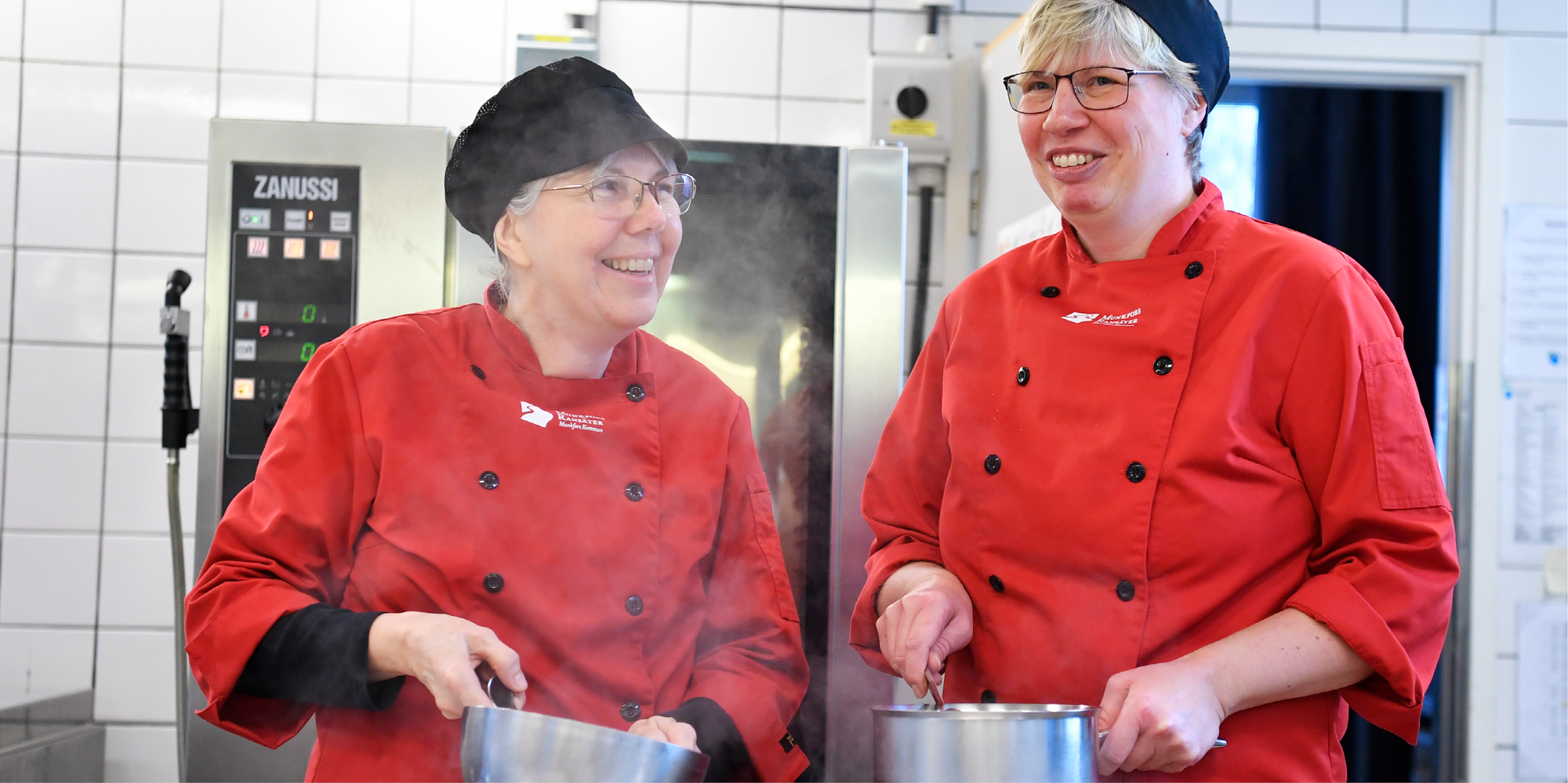 Två kvinnor i röda kockrockar ler emot varandra. I händerna har de varsin ångande kastrull. 
