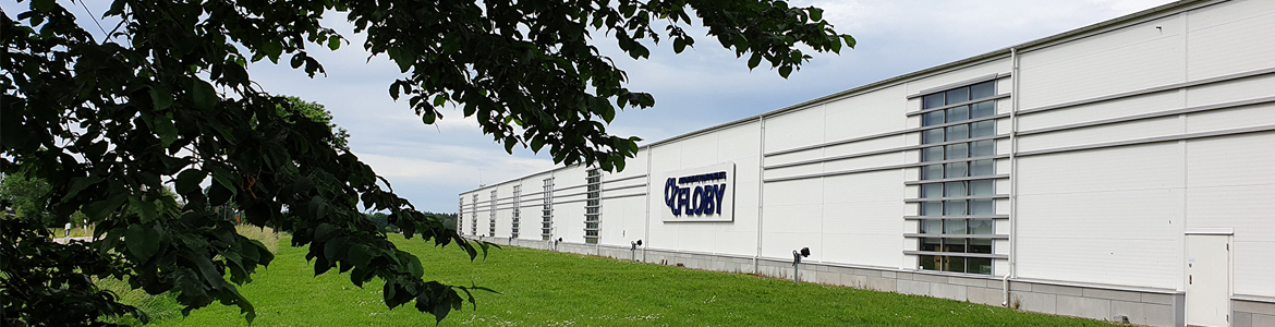 Vit lagerbyggnad med AC Floby logotyp omgiven av grönt lövverk och gräsmatta
