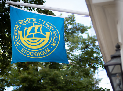 En blå flagga med gult tryck som visar en symbol med ett skepp och texten Tyska Skolan Stockholm.gga Karlavägen.jpg